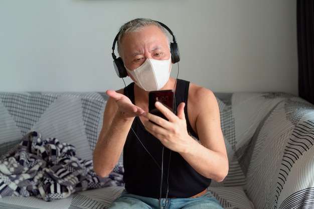 Mature Japanese man with mask video calling with headphones at home under quarantine