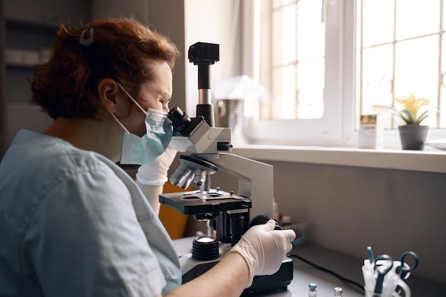 Mature lab assistant in mask looks into microscope researching sample at workplace