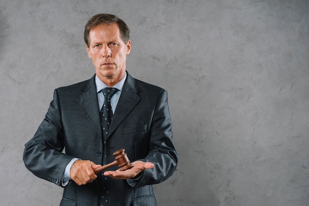 Photo mature male lawyer hitting gavel over his palm hand against gray textured backdrop