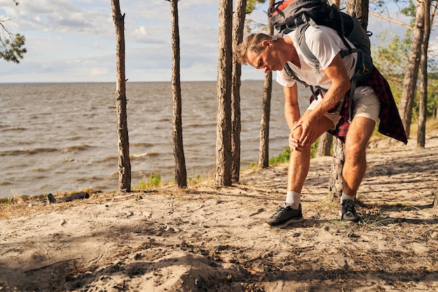 Mature male with backpack is stopping on way and suffering pain in knee while going hiking near sea shore