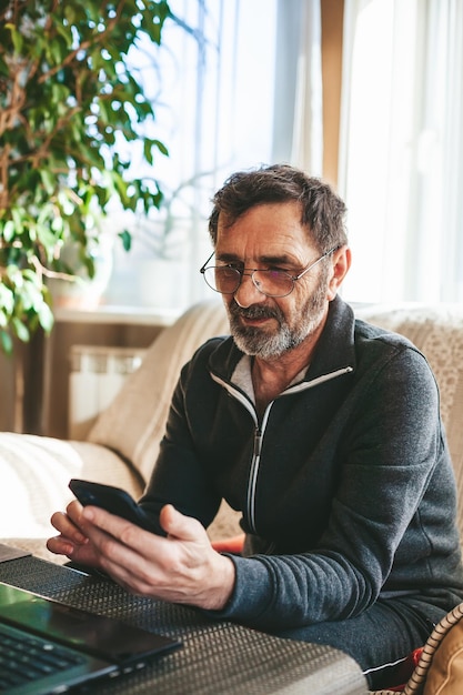 Mature man in glasses sitting on a couch texting or messaging or searching or reading on a modern smartphone Seniors using technology concept