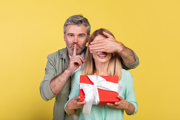 Mature man got surprise for his loving wife woman holding gift box man covering her eyes and showing silence gesture