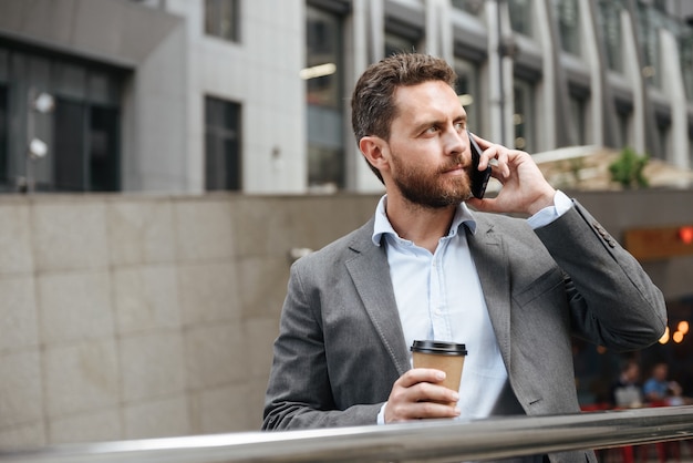 mature man in gray suit looking aside, while standing and holding takeaway coffee in front of modern business center during business call