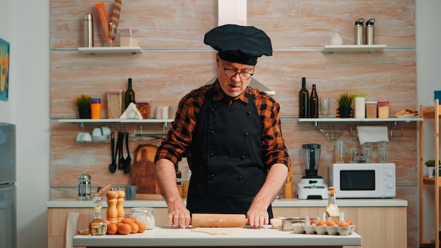 Mature man preparing homemade pizza at home kitchen. Happy elderly chef with bonete using wooden rolling pin kneading raw ingredients to baking traditional cookies, sprinkling, sieving flour on table.