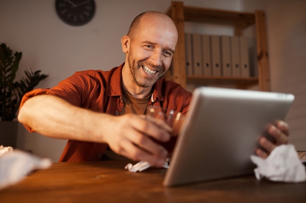 Mature man sitting at the table and using digital tablet for online conversation he talking to his friend and drinking alcohol