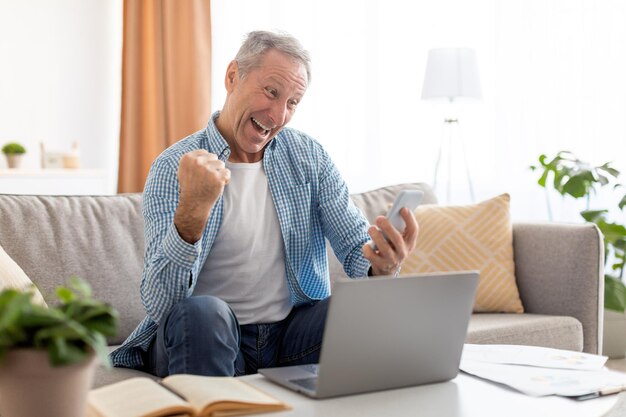 Mature man using cellphone celebrating success shaking fists screaming yes