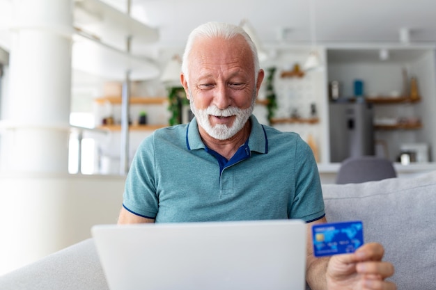 Mature man using laptop holding plastic credit or debit card senior grey haired customer making secure internet payment shopping or browsing online banking service entering information