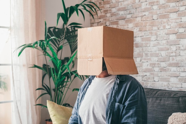 Mature man wearing empty carton box sitting on sofa in the living room of house Caucasian guy with face hidden or covered in cardboard delivery container while relaxing on couch at home