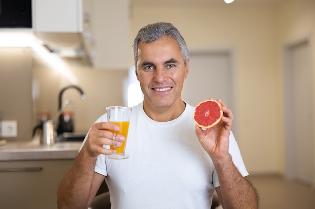 Mature man in white casual t-shirt holding half of grapefruit and glass of citrus juice and smiling