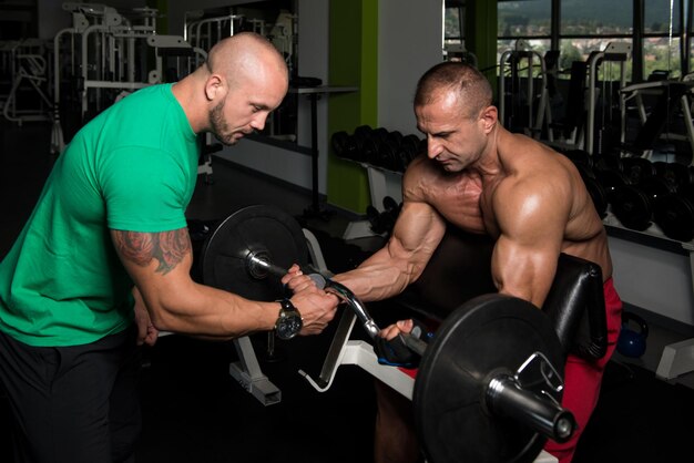 Mature Man Working Out In Gym  Doing Biceps Exercise On Machine With Help Of His Trainer