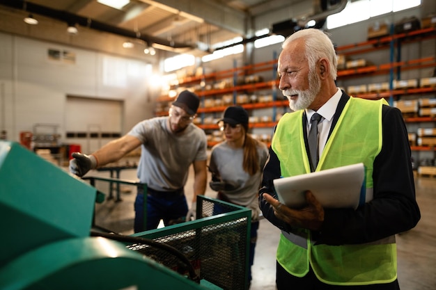 Mature manager writing notes in clipboard while overseeing his workers who are working at the machine in industrial warehouse