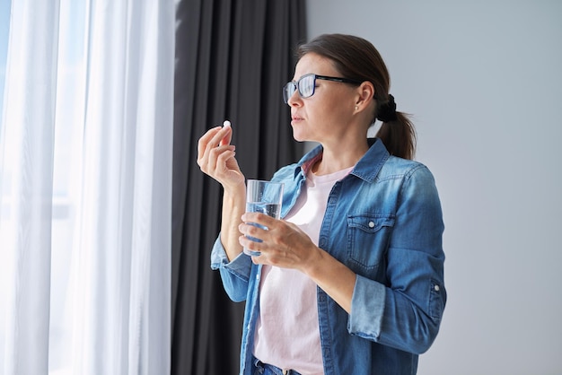 Mature middle-aged woman in casual clothes at home holding pill and glass of fresh water. Headaches, depression, medicine supplements vitamins, sedatives, menopause