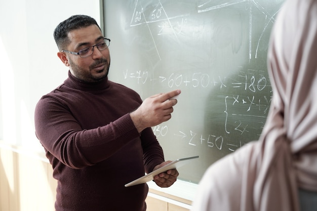 Mature mixed-race teacher explaining subject to student in hijab by blackboard