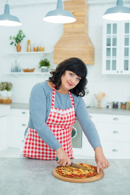 Mature pretty woman cuts readymade pizza with a knife in the kitchen Household chores and cooking