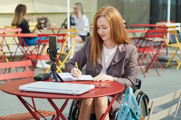 Mature redhaired woman who uses a wheelchair uses a smartphone for work remote learning makes notes in a notebook