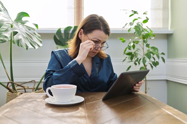 Mature serious woman at home reading mail, news, social networks on digital tablet. Morning female in pajamas with cup of tea