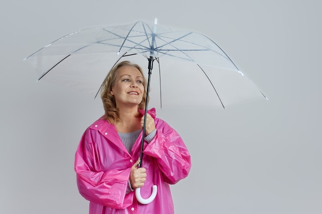 Mature smiling blonde woman in magenta raincoat with transparent umbrella on gray background