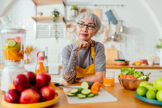 Mature smiling tattoo woman eating salad fruits and vegetables Attractive mature woman with fresh green fruit salad at home Senior woman apron standing in the kitchen counter relaxing in house