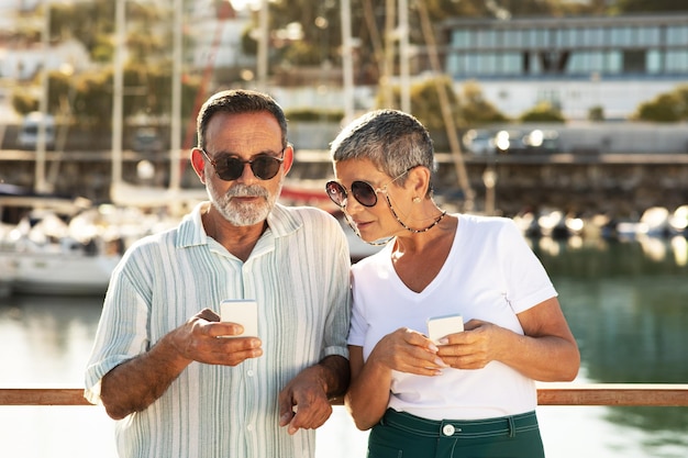 Mature spouses using phones posing at marina pier with sailboats