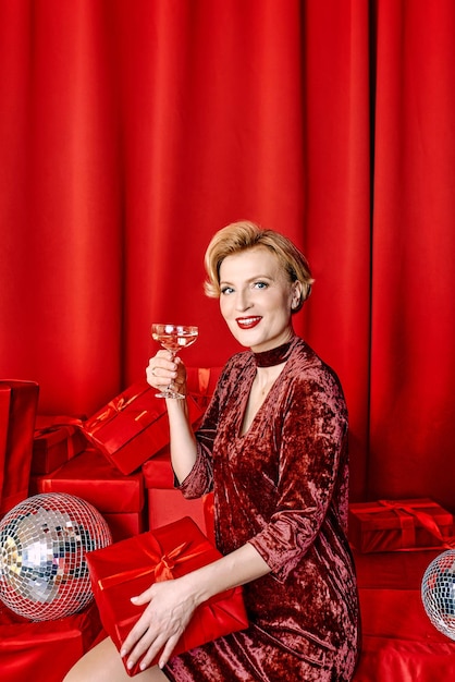 Mature stylish elegant woman with glass of sparkling wine with present on red background.