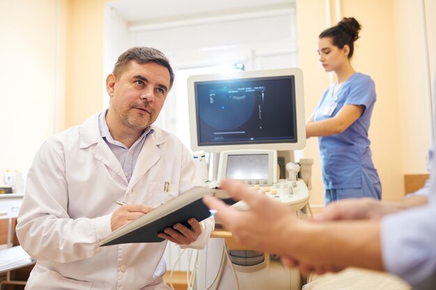 Mature ultrasound specialist in lab coat making notes in medical card while talking to patient in procedure room