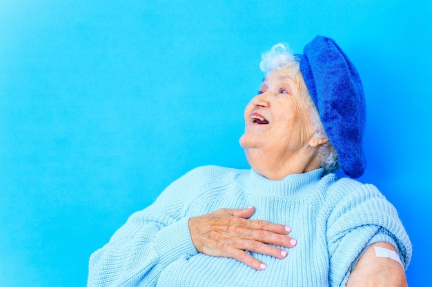 Mature woman 80 years old showing her arm with bandage after receiving vaccination in blue studio background