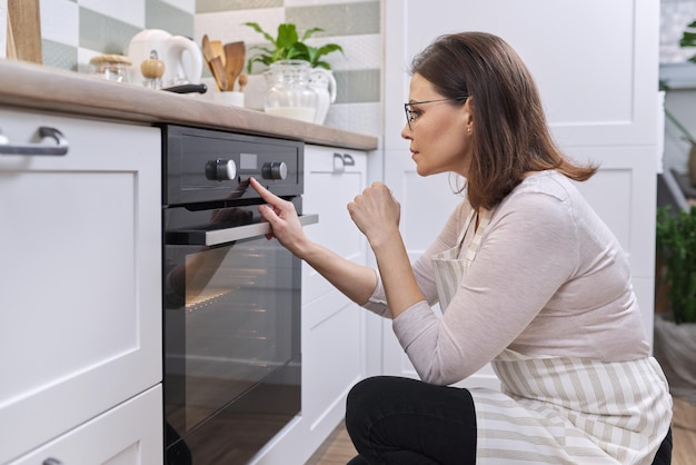 Mature woman in apron near the oven in kitchen. Housewife turning on the settings on stove, copy space