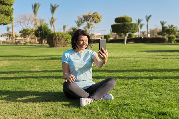 Mature woman doing morning exercises on green grass with smartphone video workout