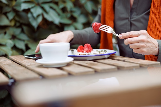 Mature woman eats toast with cream and strawberries taking mobile phone at table on outdoors