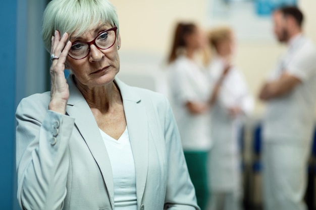 Mature woman holding her head and feeling worried while thinking of something in a hallway at clinic Team of doctors in the background