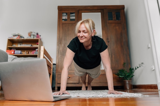 Mature woman at home doing sport yoga on the floor with laptop online classes