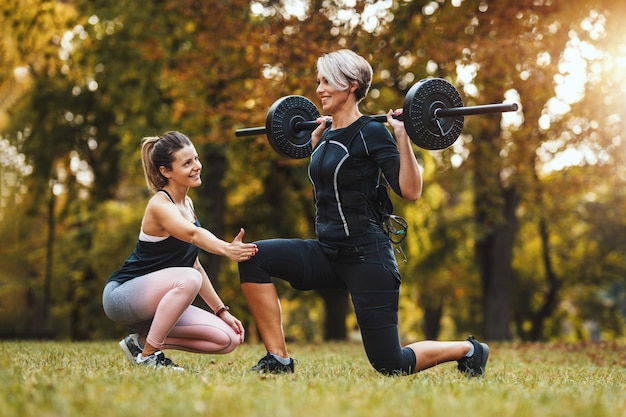 A mature woman is doing lunges exercises with personal trainer in the park, dressed in a black suit with an EMS electronic simulator to stimulate her muscles.