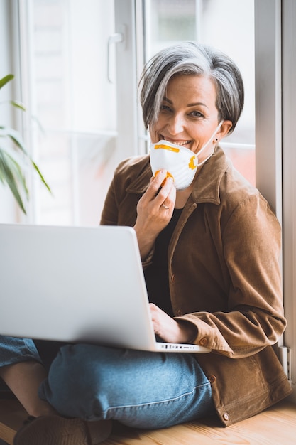 Mature woman lowering fp1 mask and smile looking at camera while works laptop computer