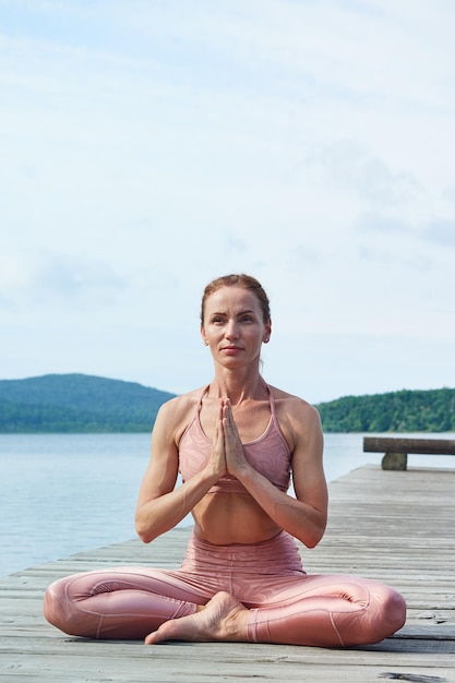 Mature woman in a pink tracksuit sits in a lotus position practices yoga near the sea Healthy lifestyle concept