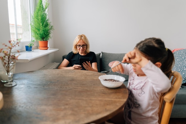 Mature woman reading tablet or ebook while preschool girl eating alone Social media addiction