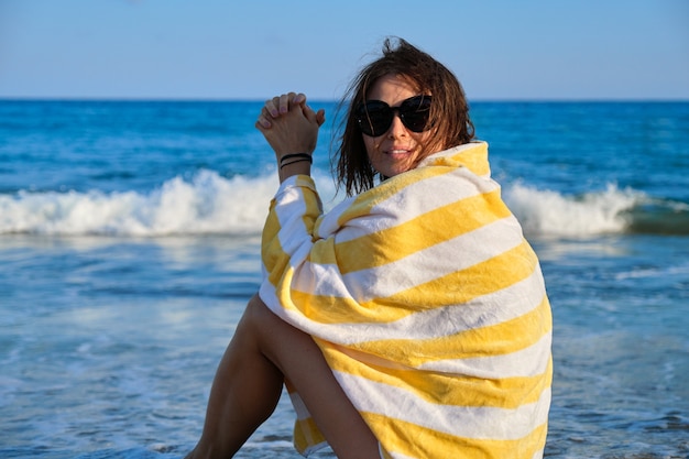 Mature woman sitting on seashore resting beautiful female with beach towel on her shoulders. Beautiful sunset scenic seascape background. Relax, leisure, beauty, ocean, lifestyle of middle-aged people