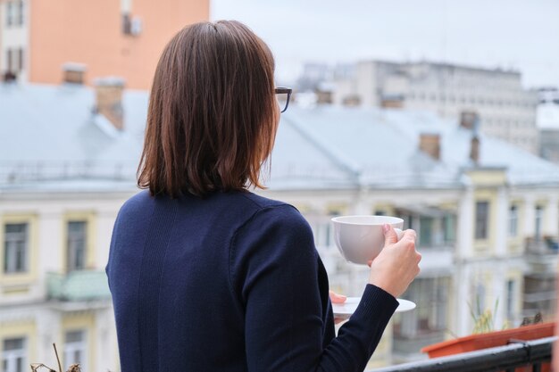 Mature woman standing on open balcony with cup of tea, female looks at city while in isolation, quarantined during viral infection, copy space