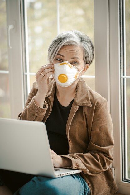 Mature woman straightens the mask works laptop computer sitting on a windowsill.