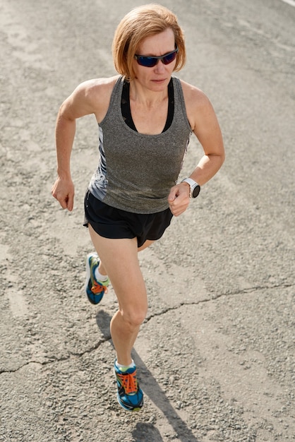 Mature woman in sunglasses runner running on country road in the morning