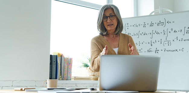 Mature woman teaching mathematics while standing near the whiteboard and looking at laptop