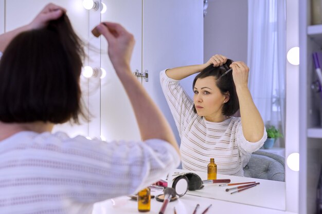 Mature woman treating caring for hair using cosmetic medical products in bottle with pipette. Middle-aged female sitting at home looking in mirror. Hair loss problem, special professional preparations