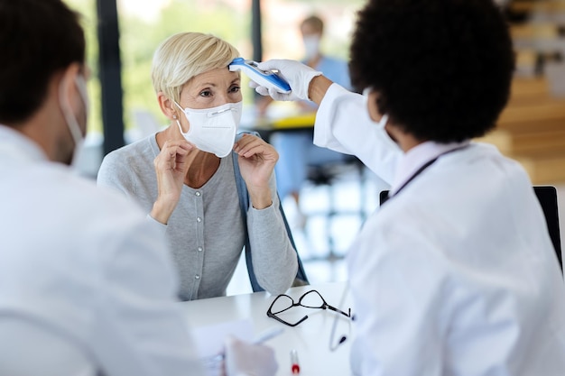 Mature woman with face mask getting her temperature measured during medical appointment