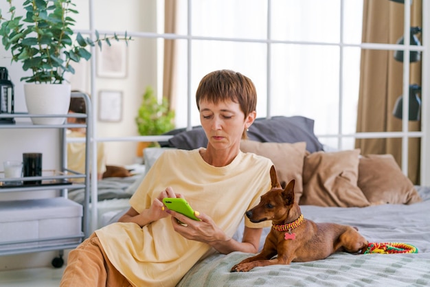 Mature woman with a phone in her hand with a pygmy pinscher in the bedroom