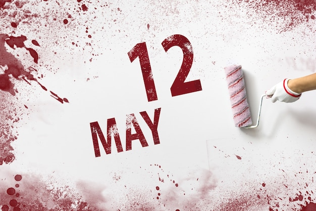 May 12nd. Day 12 of month, Calendar date. The hand holds a roller with red paint and writes a calendar date on a white background. Spring month, day of the year concept.