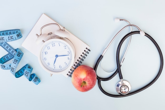 Measuring tape; alarm clock; spiral notepad; apple and stethoscope on blue background