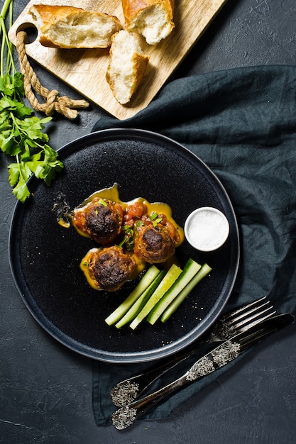 Meat balls in tomato sauce on a black plate, cucumbers, salt, bread. 