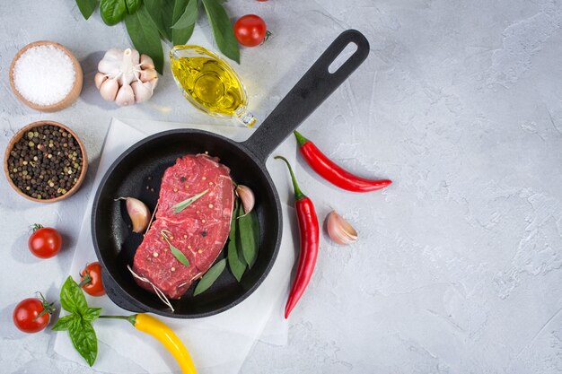 Meat steak on frying pan with ingredients for cooking on a gray concrete background Top view. Copy space.