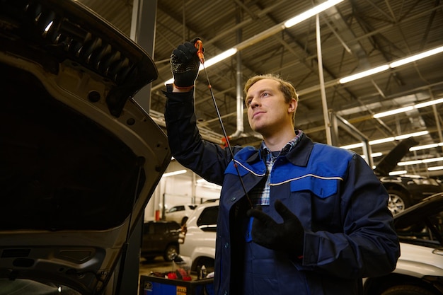 Mechanic in garage examining the car engine oil dipstick checking motor oil level at a car. Car engineer technician in the car repair shop. Automobile maintenance and auto service concept