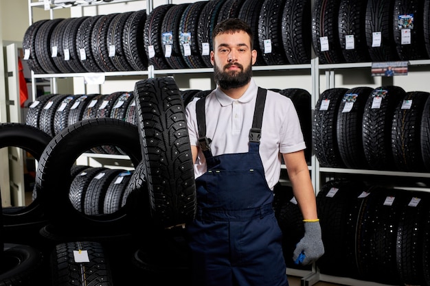 Mechanic holding a tire and showing wheel tires at car repair service and auto store shop, young bearded male in uniform working in automobile repair garage