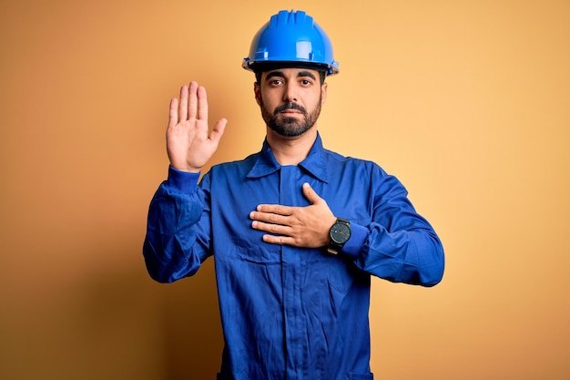 Mechanic man with beard wearing blue uniform and safety helmet over yellow background Swearing with hand on chest and open palm making a loyalty promise oath